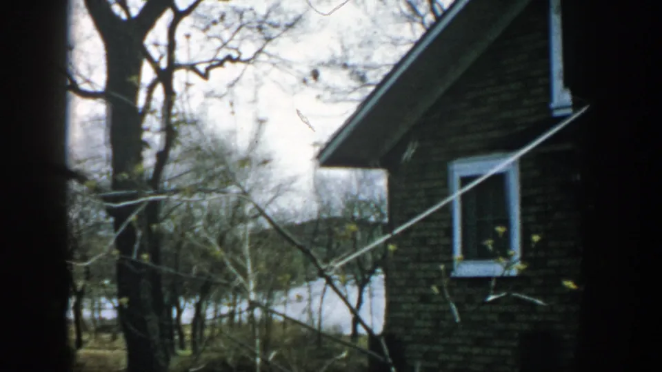 1957 Wisconsin 8mm frame of a brick house corner near leafless trees, with a white-trimmed window and open ground in the distance.