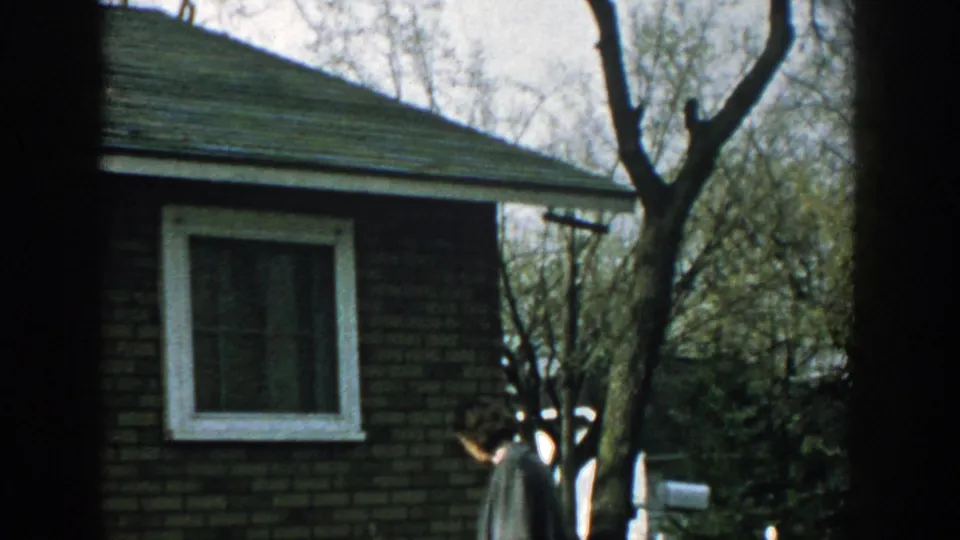 1957 Wisconsin home-movie frame with a brick house, white-trimmed window, bare trees, and a woman at the edge of the yard.
