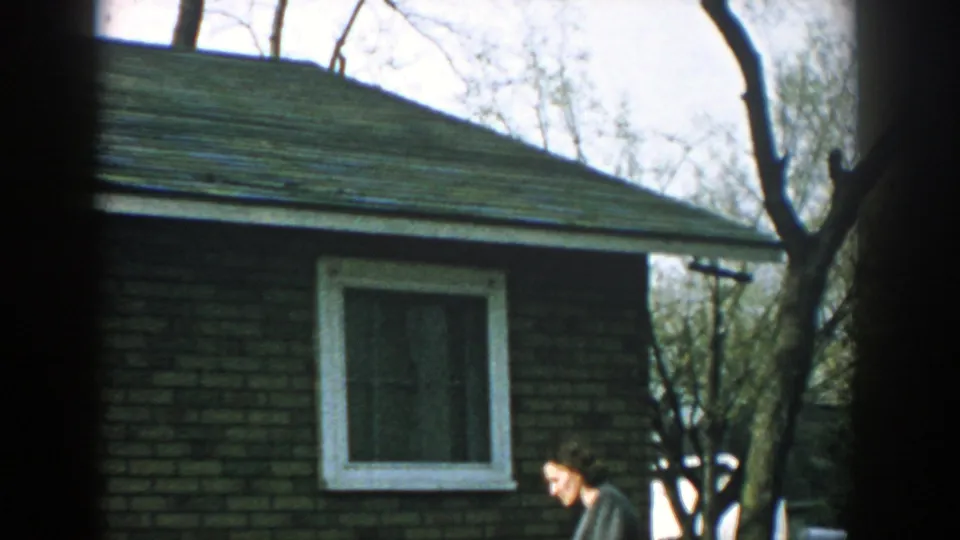 1957 Wisconsin frame showing a woman in a coat moving beside a brick house with a white-trimmed window and bare trees.