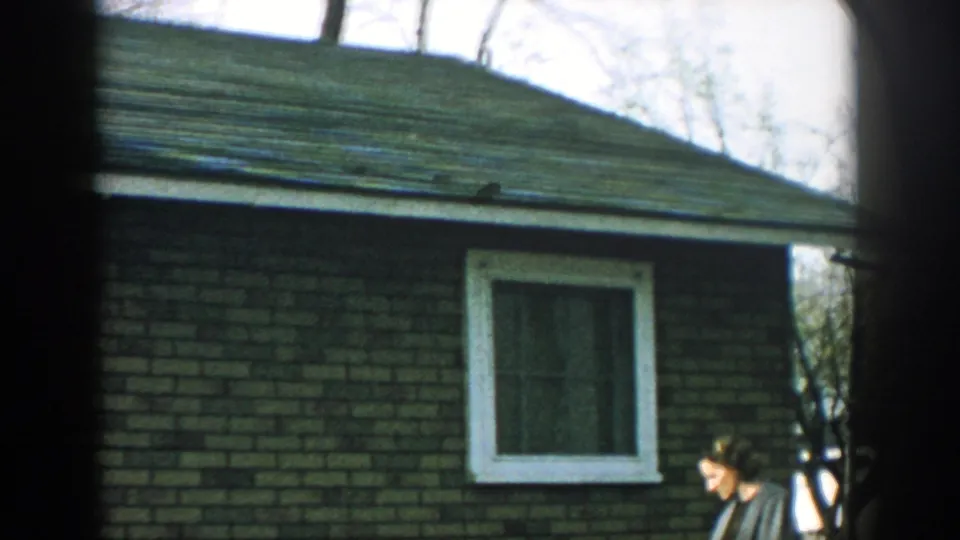 1957 Wisconsin home-movie frame of a woman near a brick house exterior, white-trimmed window, yard, and leafless trees.