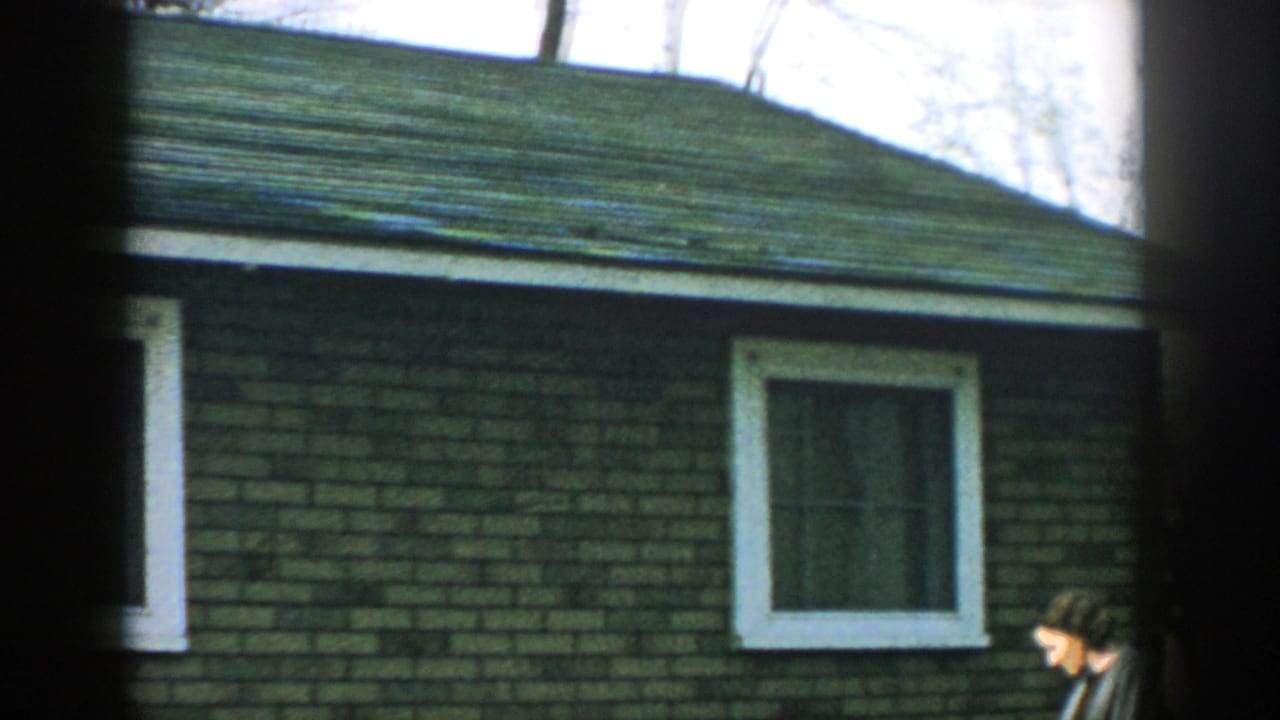 1957 Wisconsin 8mm frame showing a brick residential exterior with white window trim and a green shingled roof, partly obscured by dark foreground framing.