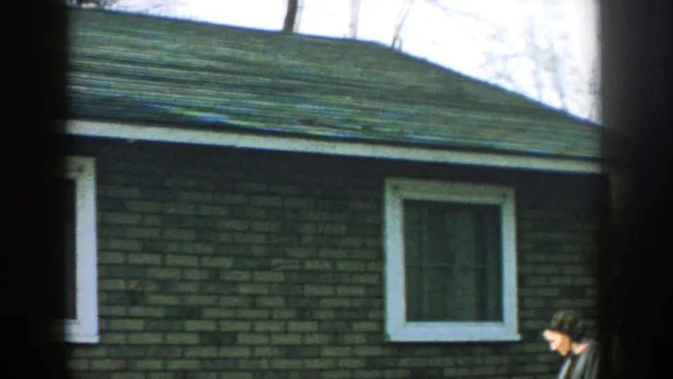 1957 Wisconsin 8mm frame showing a brick residential exterior with white window trim and a green shingled roof, partly obscured by dark foreground framing.