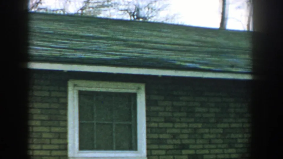 1957 Wisconsin home-movie frame of a brick residential building with a white-trimmed window and a briefly visible adult woman near the lower edge.