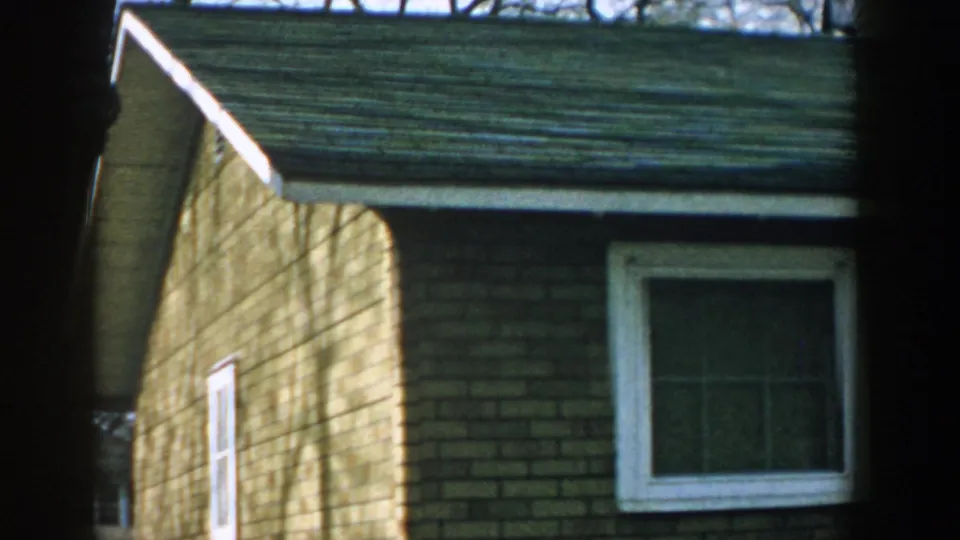 1957 Wisconsin 8mm view of a brick residential side wall, white trim, and green roofline, with dark bars crossing the foreground.