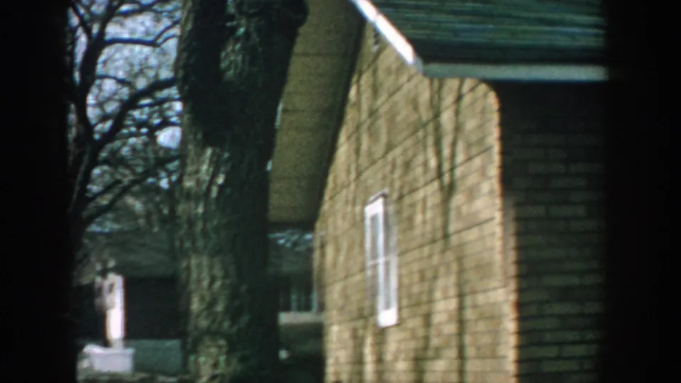 1957 Wisconsin home-movie frame showing brick residential buildings, white-trimmed windows, a green roof, and a large foreground tree trunk.