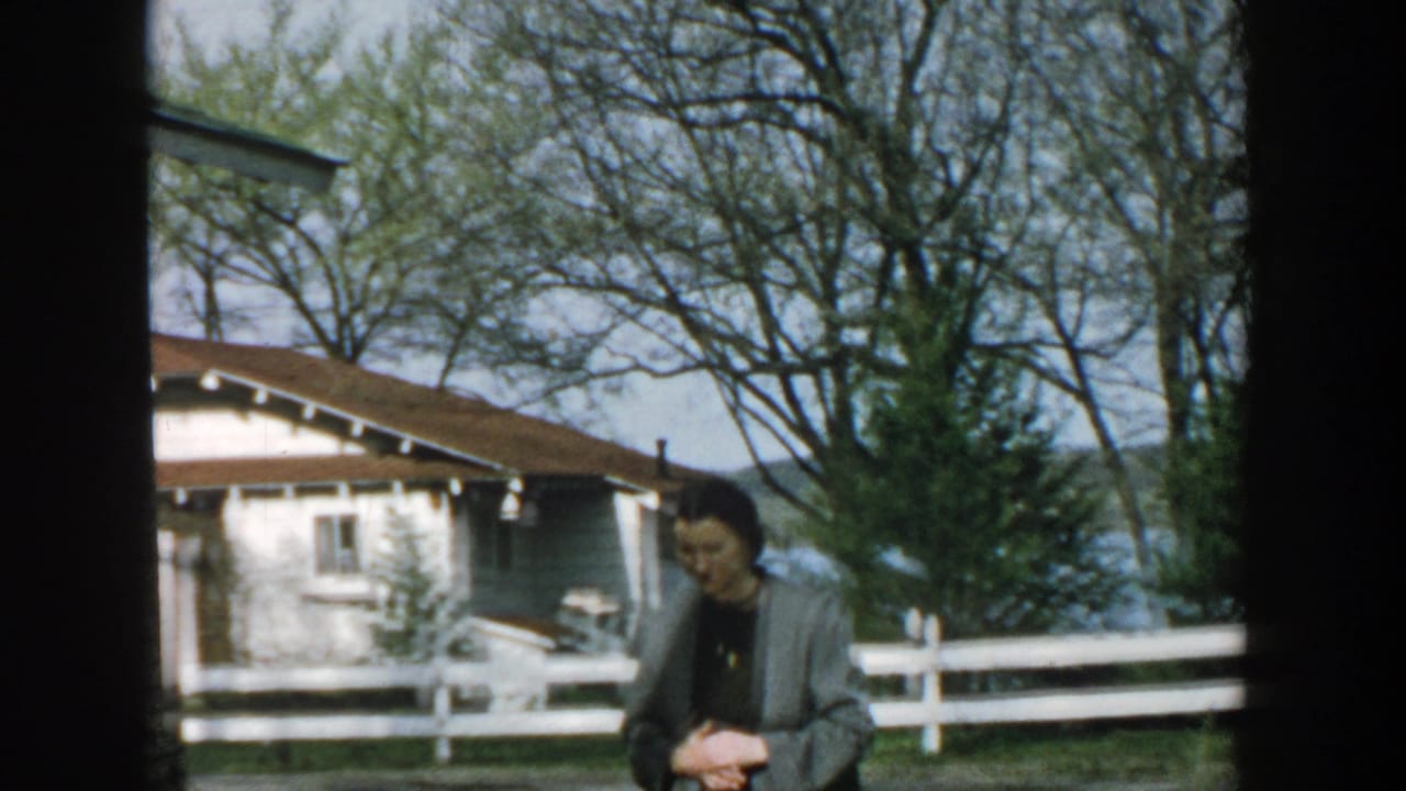 Adult woman in a yard near a white fence, cottages, trees, and water.