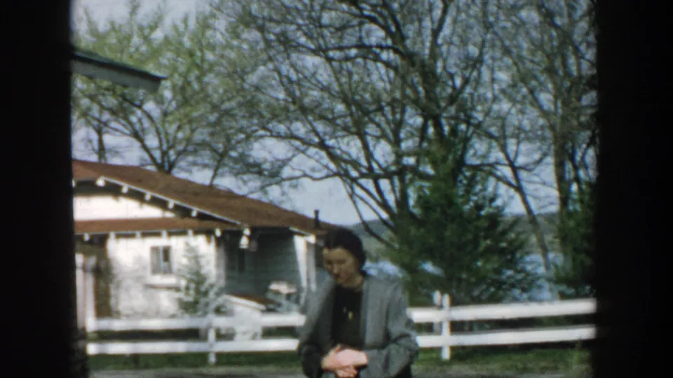In 1957 Wisconsin home-movie footage, an adult woman stands in a rural yard with cottages, a white fence, trees, and distant water behind her.