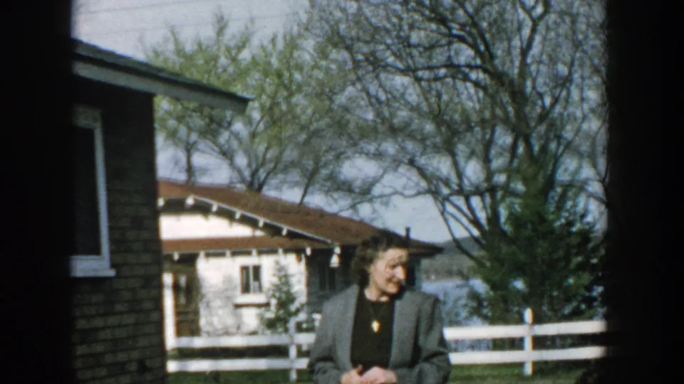 An adult woman smiles near a brick building in a Wisconsin yard, with a white fence, cottage roofs, trees, and water in the background.
