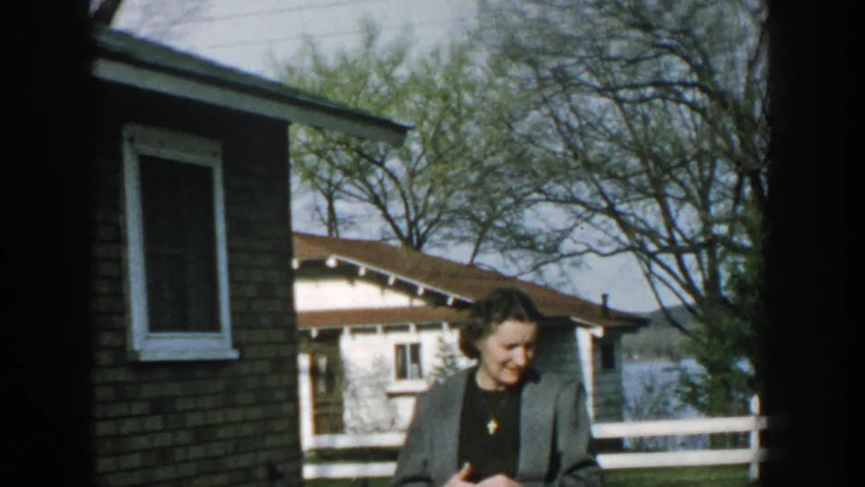 The woman looks down while standing near a brick building, with a white fence, trees, cottage, and lakeside background visible in the 1957 Wisconsin footage.