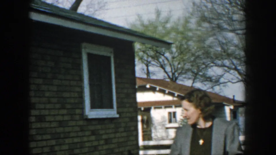 An adult woman in profile stands near a brick building in a rural Wisconsin yard, with cottages, a white fence, trees, and water beyond.
