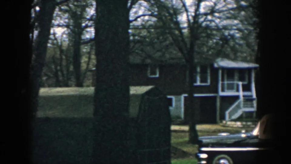 A Wisconsin residential exterior appears through trees, with white trim and part of a 1950s sedan in the driveway.