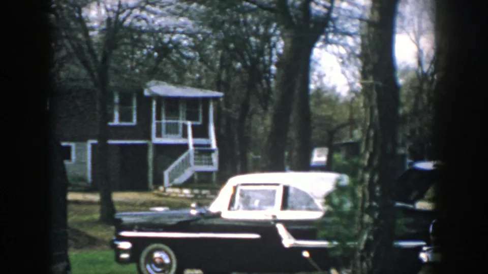Mid-century sedan parked near a wooded Wisconsin house, seen in soft 8mm color with porch stairs behind it.