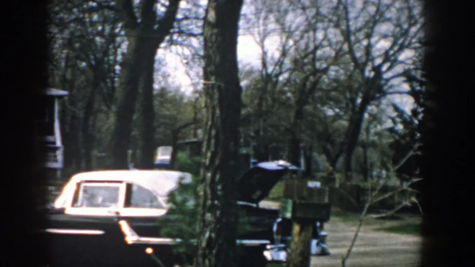 The pan settles on the sedan and wooded driveway area, with a house edge and mailbox-like post visible in the 1957 home movie frame.