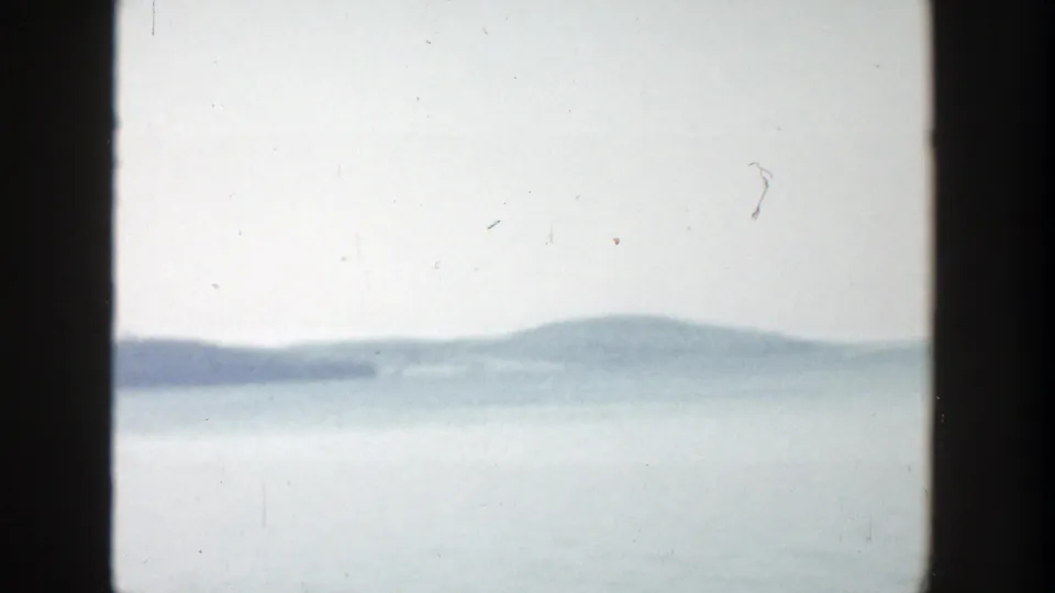 1957 Wisconsin frame with calm water, a hazy wooded shoreline, and soft 8mm film grain.