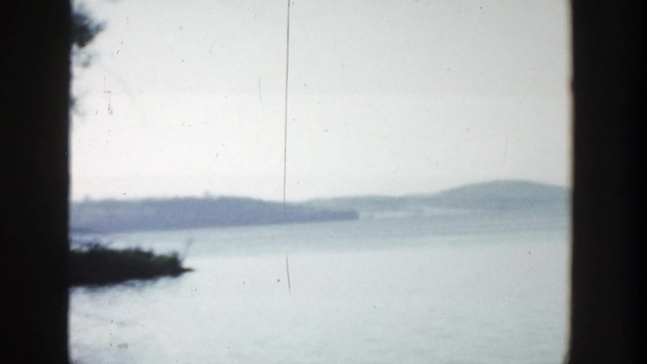 Calm lake water seen between dark vertical borders with distant wooded shoreline and pale sky.