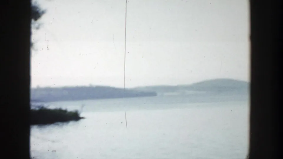 1957 Wisconsin 8mm frame showing calm lake water, distant tree-lined shore, pale sky, and dark vertical film or window borders.