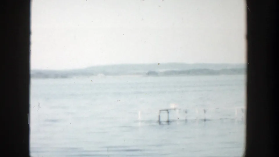 1957 Wisconsin lake view with open water, a hazy distant shore, dark frame edge, and a small dock or swim platform at right.