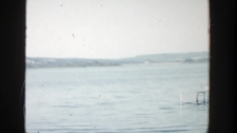 1957 Wisconsin 8mm frame of a broad lake surface, distant low shoreline, pale sky, and heavy dark vertical framing edges.