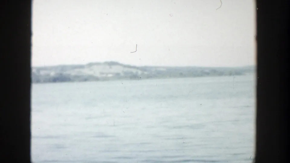 1957 Wisconsin 8mm frame showing calm open water, a distant tree-covered shoreline, low hills, and pale overcast sky within dark edge framing.