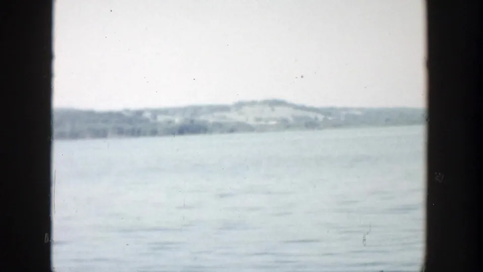 1957 Wisconsin 8mm frame with a broad lake view, distant low hills, muted blue-gray water, pale sky, and visible dark vertical framing.