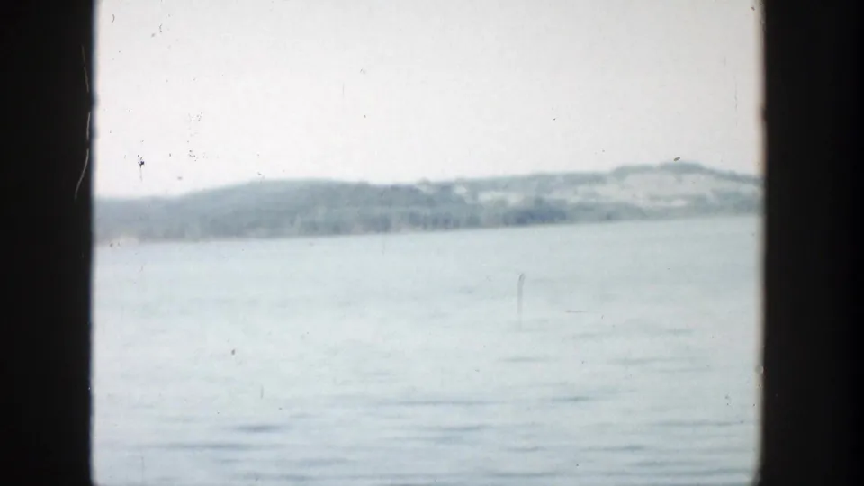 1957 Wisconsin 8mm frame showing calm water in the foreground, a distant wooded shoreline, low hill shapes, pale sky, and visible dust marks.