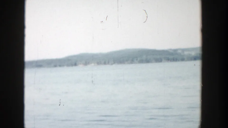 1957 Wisconsin 8mm frame with blue-gray water, a far tree line, low rolling hills, pale sky, scratches, and dark edge framing.