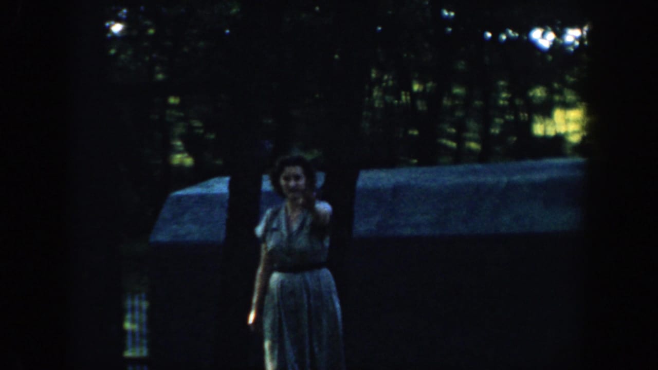 1957 Wisconsin 8mm frame of a woman in a light dress facing the camera in a shaded wooded suburban area.