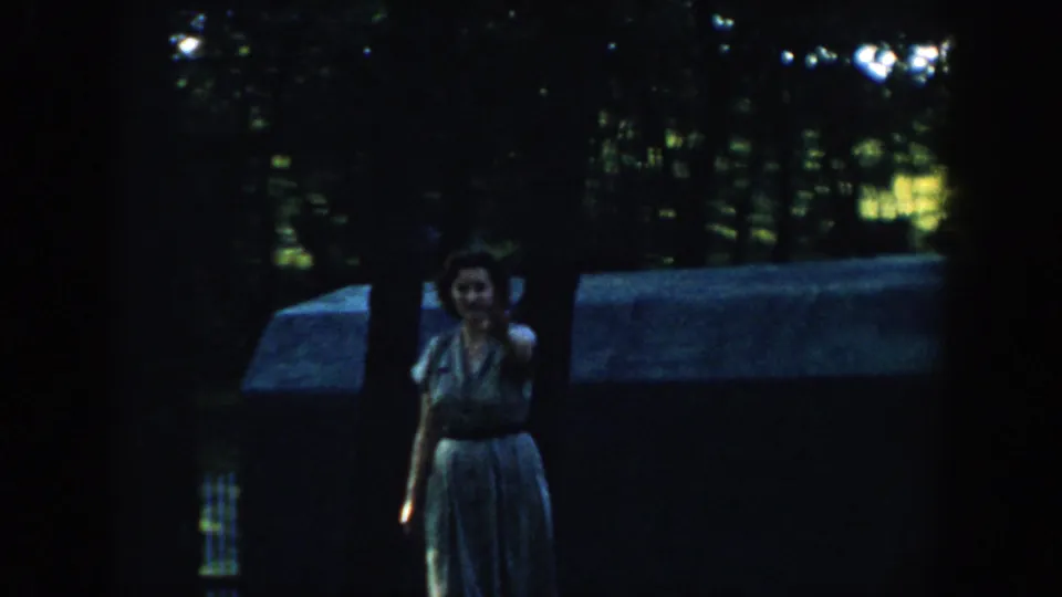 1957 Wisconsin 8mm frame of a woman in a light dress facing the camera in a shaded wooded suburban area.