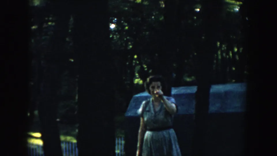 1957 Wisconsin home-movie frame showing a woman in a light dress pointing toward the lens in a dark wooded setting.