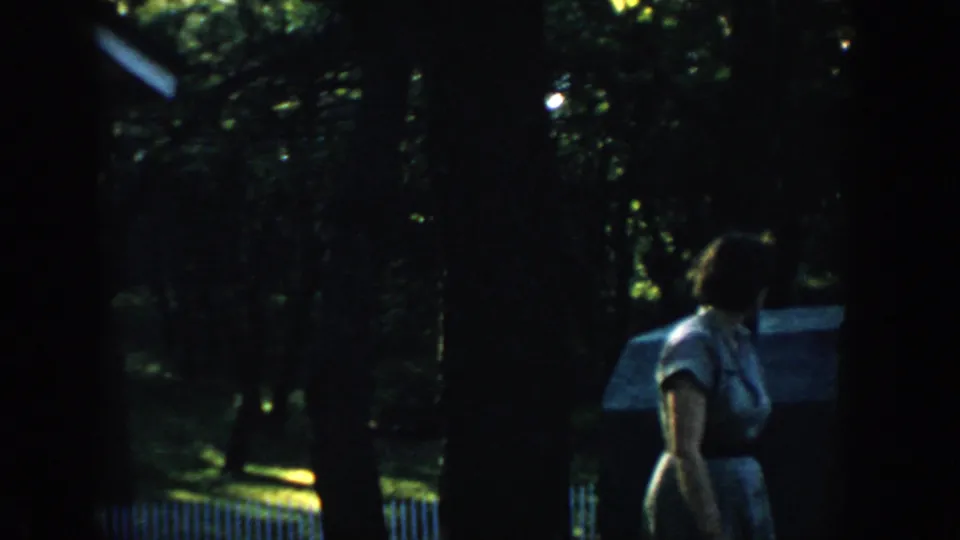 1957 Wisconsin 8mm frame of the woman turned in profile while moving through a shaded outdoor residential area.