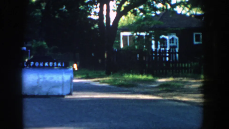 1957 Wisconsin home-movie view of a mailbox by a shaded suburban street with trees, a fence, and a house beyond it.