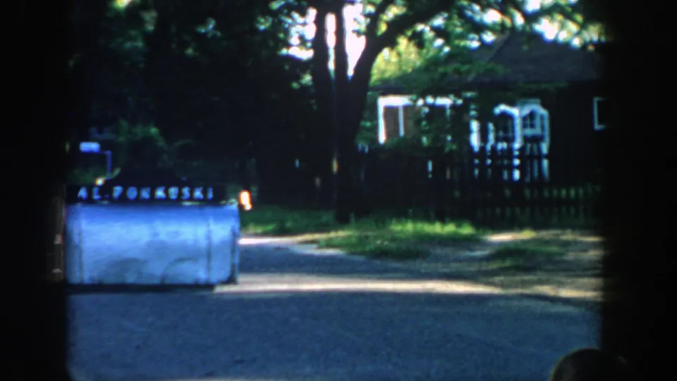 Soft 8mm frame from 1957 Wisconsin showing a roadside mailbox, quiet street, dark fence, leafy trees, and a modest house.