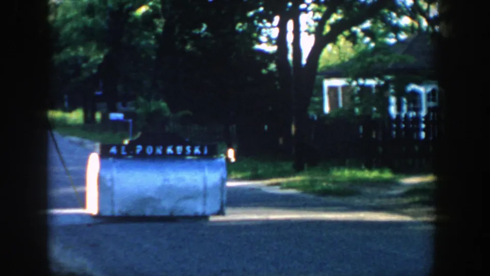 1957 Wisconsin 8mm frame with the 42 PONKUSKI mailbox at roadside, backed by trees, a fence, a driveway, and a house.