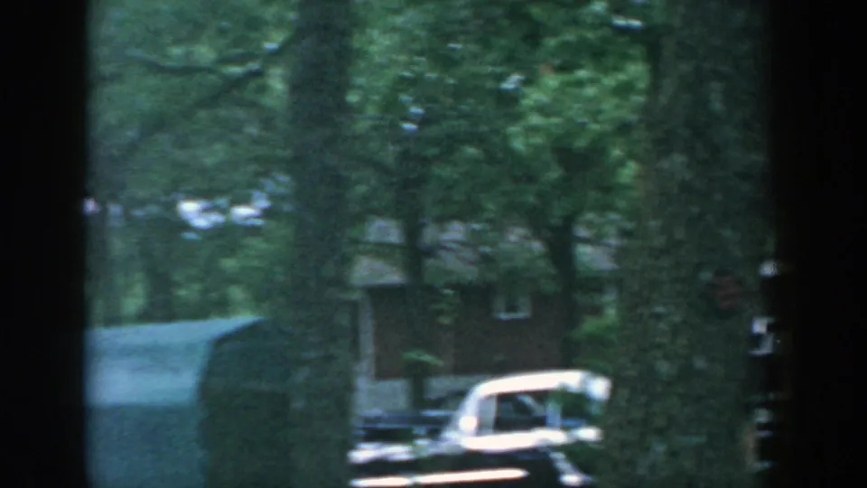 1957 Wisconsin home-movie frame with trees, a parked mid-century car, and a building partly obscured by dark window framing.