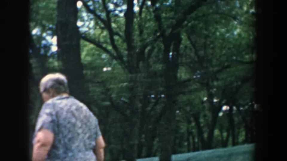 1957 Wisconsin frame showing a woman in a blue patterned dress walking through a leafy outdoor area beside dark window framing.