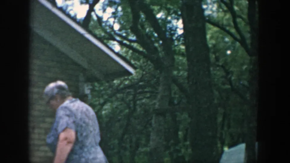 1957 Wisconsin home-movie frame of a woman in a blue patterned dress walking near a tan stone building under trees.