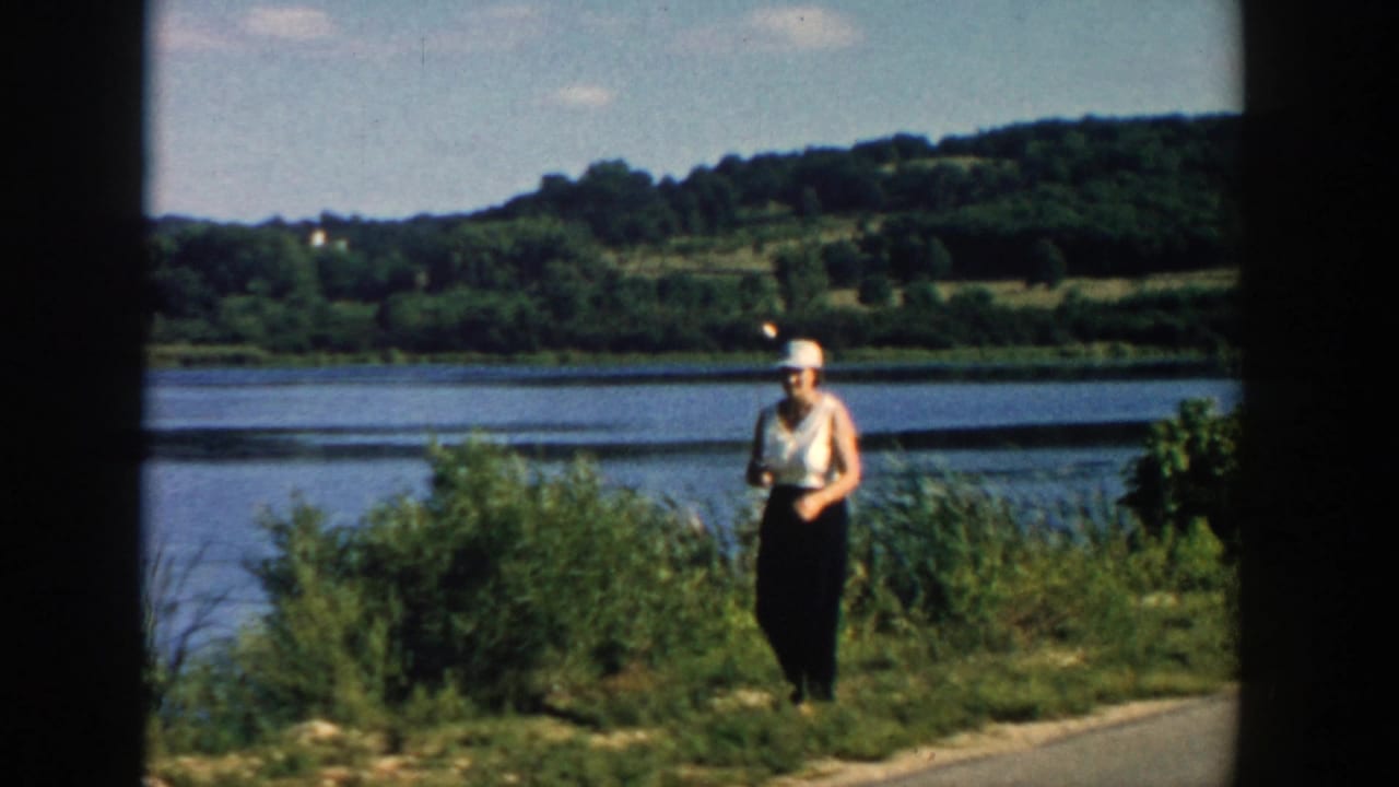 Woman in a hat stands by a grassy riverbank holding a fishing rod.