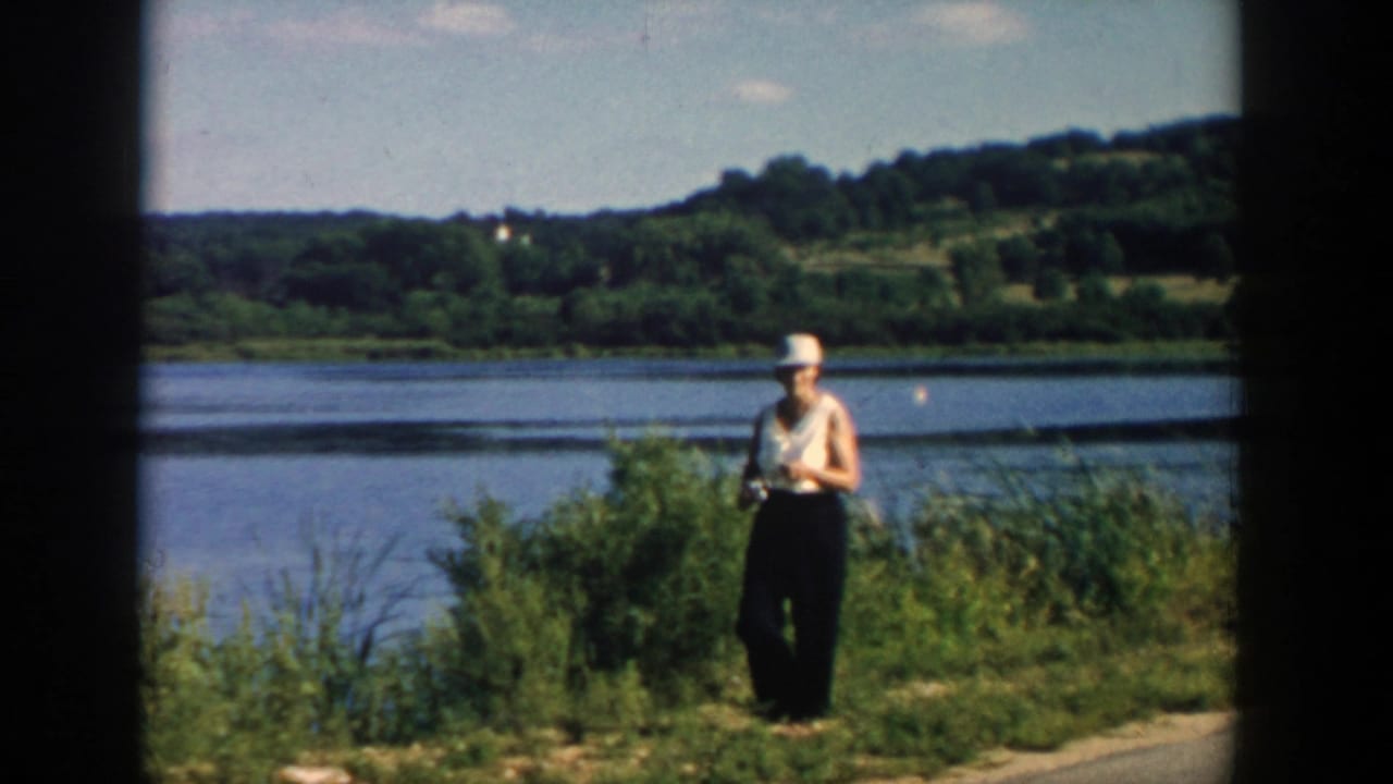 Woman in a hat holds a fishing rod along a Wisconsin riverside, with calm water and green hills in the background.
