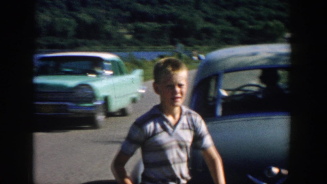 A boy is seen near the road with 1950s cars passing, while the river and wooded landscape remain behind him.