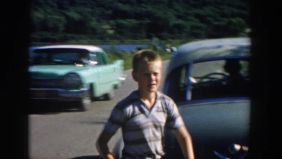 A boy is seen near the road with 1950s cars passing, while the river and wooded landscape remain behind him.
