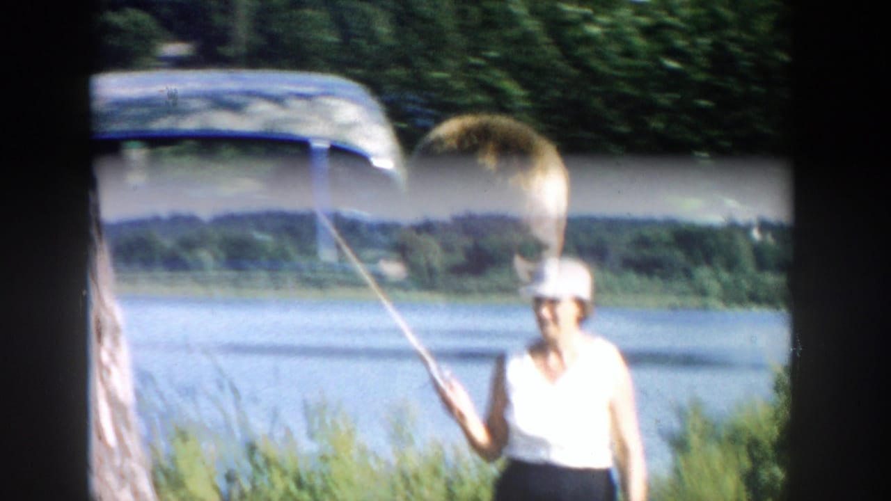 Woman in a white top casts a fishing rod beside the Wisconsin river, with a boy partially visible in the foreground.