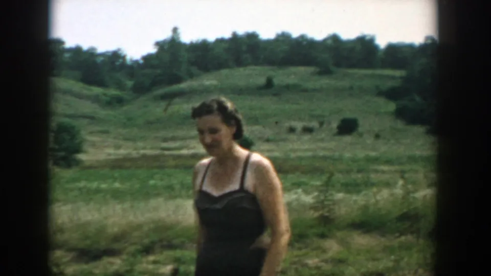 Woman in dark swimwear walks through a Wisconsin field, with rolling grass, trees, and distant cattle-like shapes behind her.