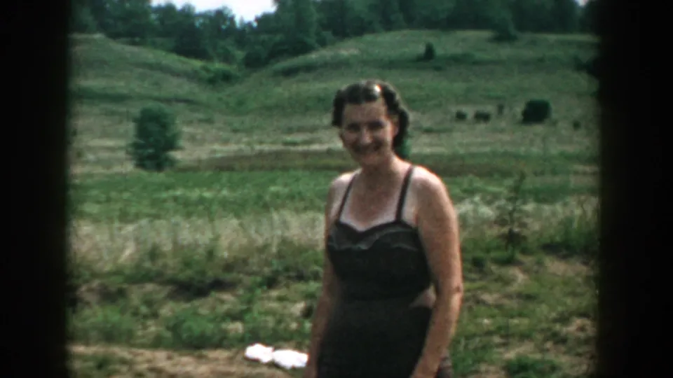 Woman in dark swimwear smiles at the camera in a 1957 Wisconsin home-movie frame, with fields and trees behind her.