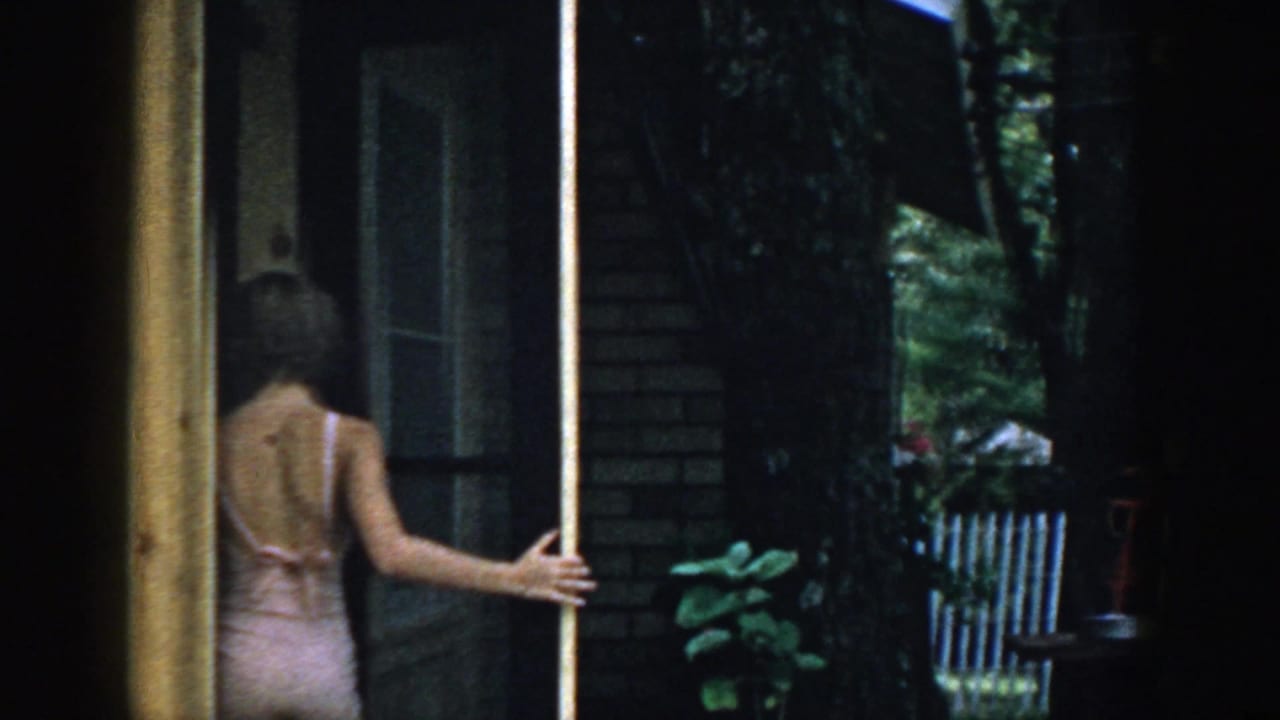 Woman in a light swimsuit opening a screen door beside a brick house.