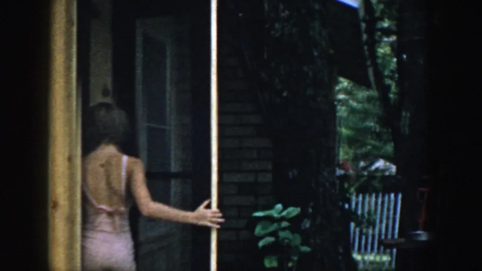 1957 Wisconsin home-movie frame of a woman in a light swimsuit at a screen door near a brick house and trees.