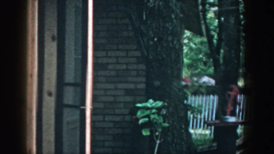 Wooded residential yard in Wisconsin with a brick house wall, window, trees, and fence visible in a 1957 home movie frame.