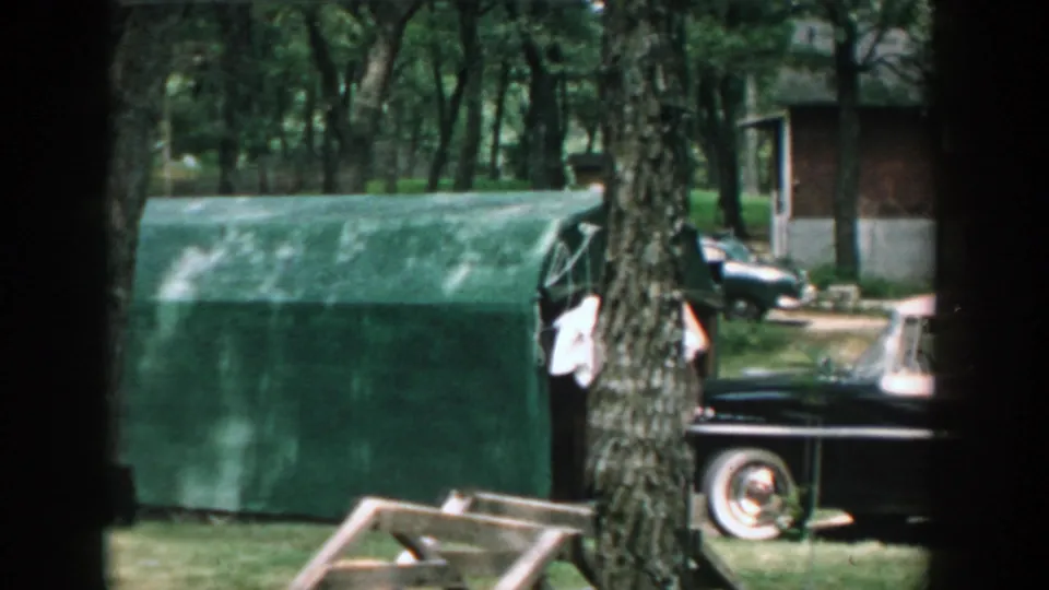 1957 Wisconsin yard view with a green tarp-covered structure, tree trunks, grass, and a classic black car in the background.