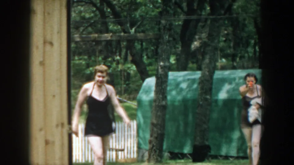 1957 Wisconsin backyard frame with one woman walking toward the camera and another holding a towel near a green covered structure.