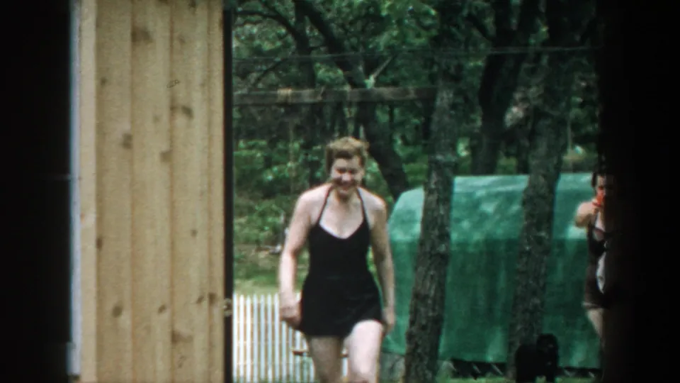 1957 Wisconsin home-movie frame showing a smiling woman in a dark swimsuit walking near trees, fence, and a green covered structure.