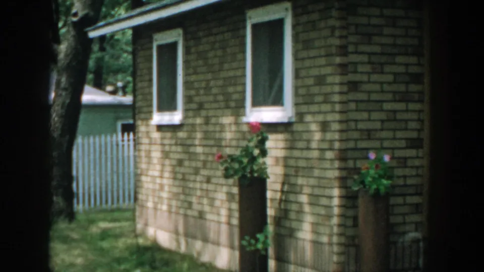 1957 Wisconsin yard frame showing a brick outbuilding, windows, flower planters, trees, and a white picket fence.